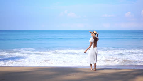 asian woman standing on the beach by the sea water when big foamy tides roll on sandy beach in summer wearing a white sundress and straw hat - slow-motion static shot