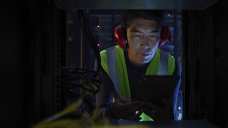 asian male it technician wearing headphones and using tablet checking computer server