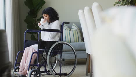 Happy-biracial-disabled-woman-in-wheelchair-drinking-coffee-and-looking-out-of-window-in-living-room