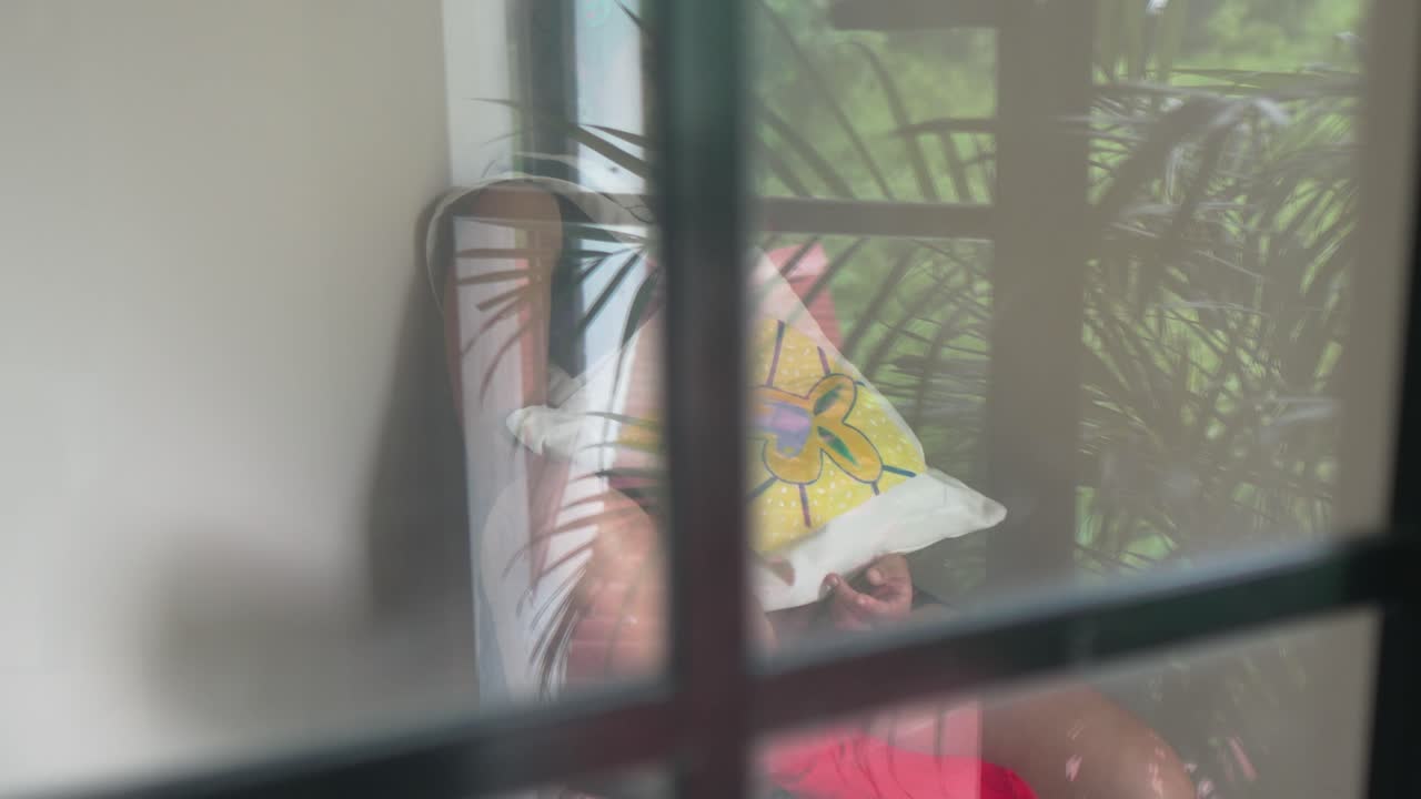 Giovane donna che si rilassa al chiuso vicino a una finestra, con in mano una borsa tote artistica colorata, con luce naturale e piante che creano un'atmosfera di stile di vita calma.