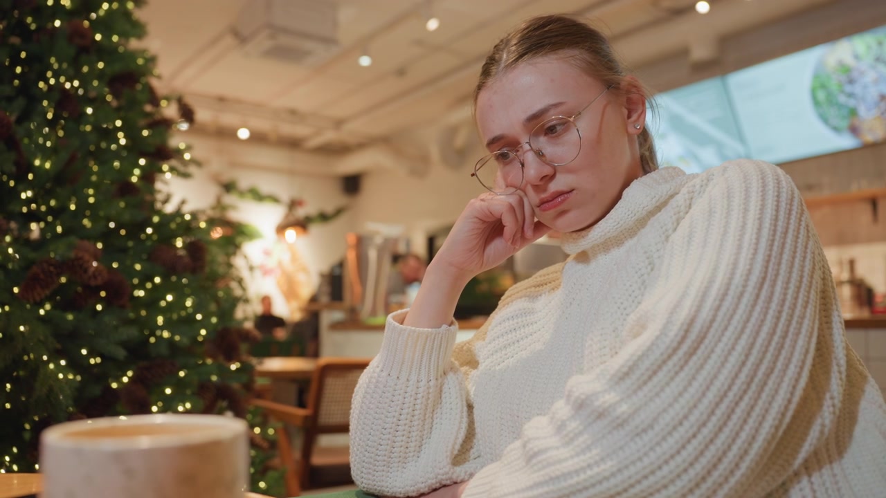 Pensive Young Woman in Glasses Sitting in a Cozy Cafe During Christmas