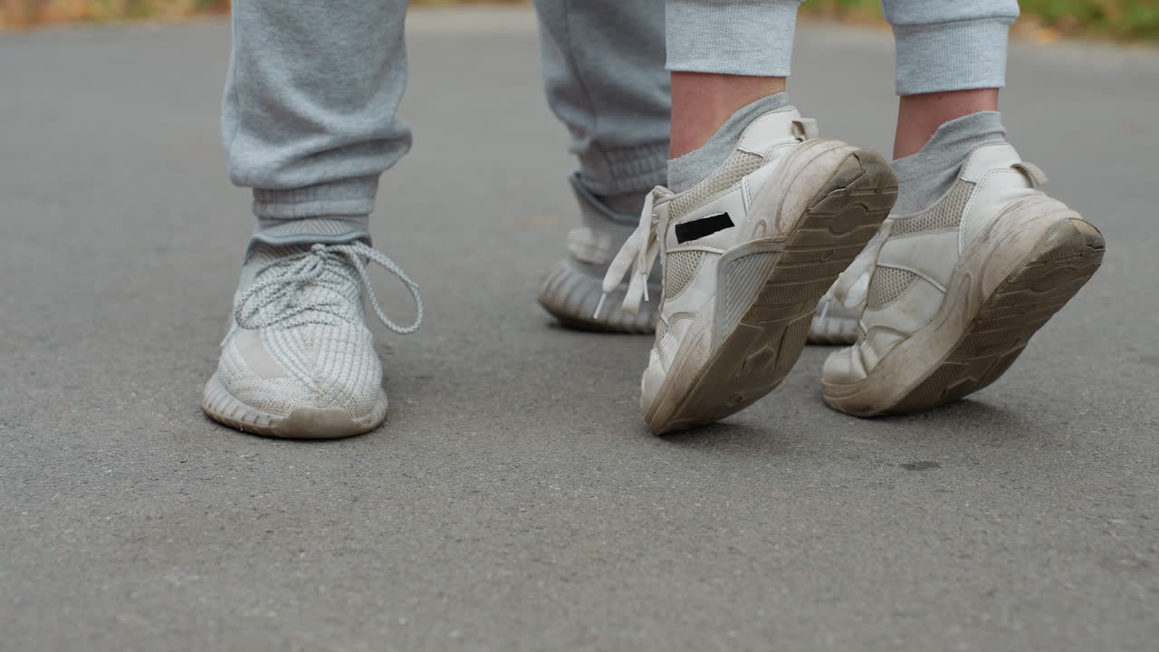 Leg view of two people wearing joggers and sneakers standing on pavement as one person stretches up on toes, raising legs, suggesting intimate moment or playful interaction outdoors in casual setting