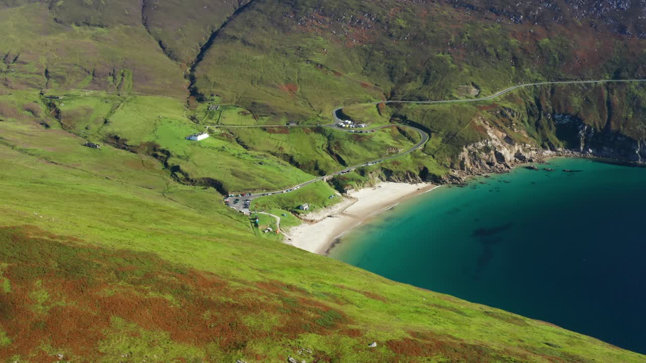 Aerial orbit of Keem Beach with calm blue Atlantic waters, cars parked by the coastline, and a winding mountain road in County Mayo