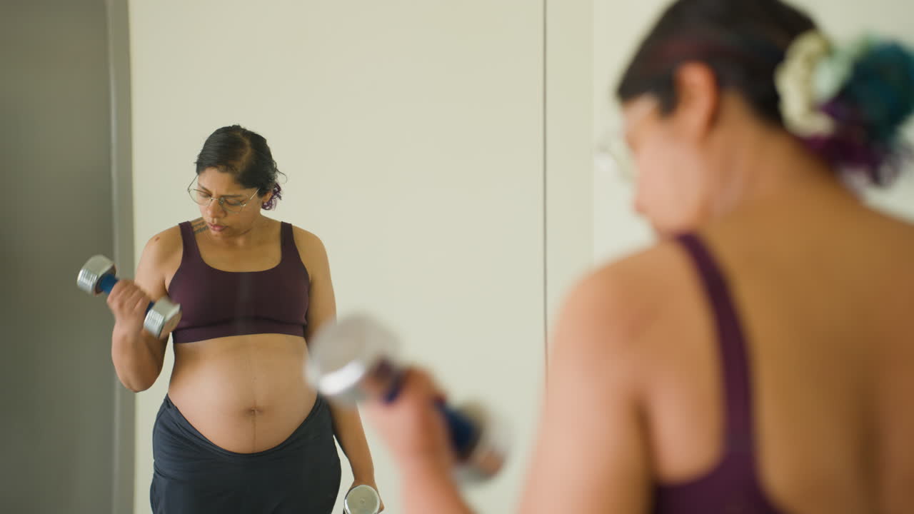 Donna indiana al terzo trimestre di gravidanza esegue esercizi di curl per bicipiti con manubri, riflessa nello specchio della palestra.