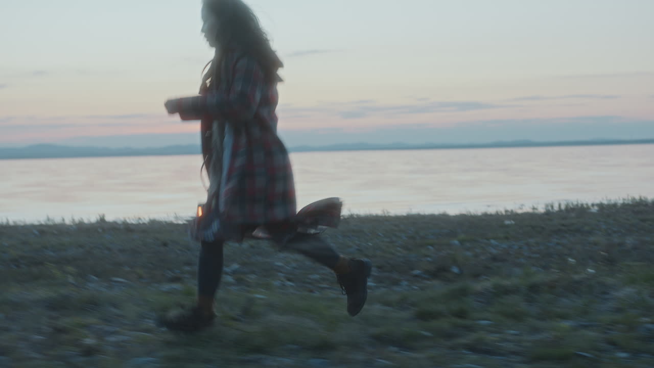 Young Woman Holding Glowing Lantern, Running along Lakeside at Dusk