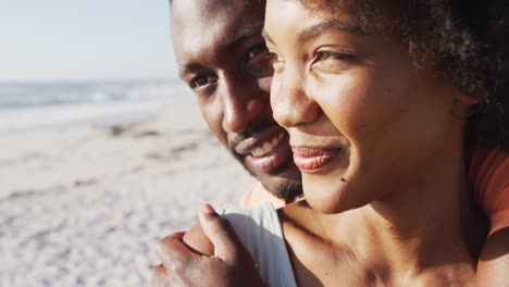 smiling african american couple embracing and looking away on sunny beach