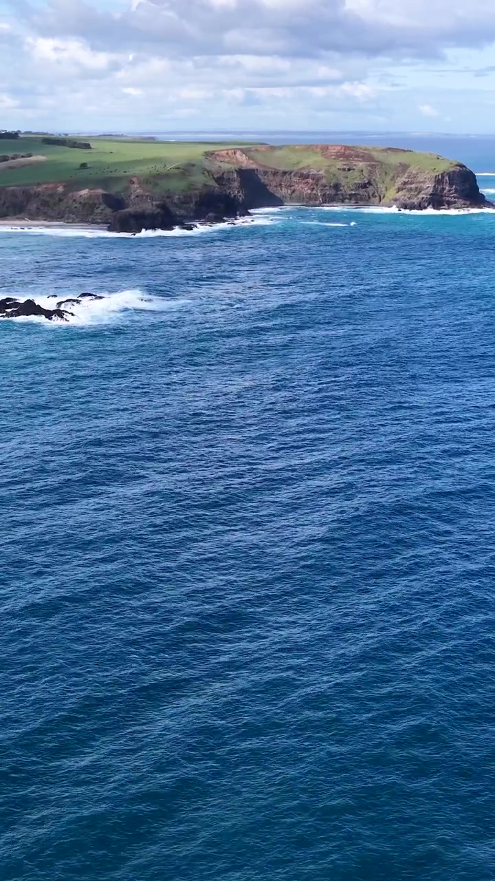 Il drone panoramica sull'oceano blu verso scogliere rocciose e un promontorio erboso sotto nuvole luminose e sparse.