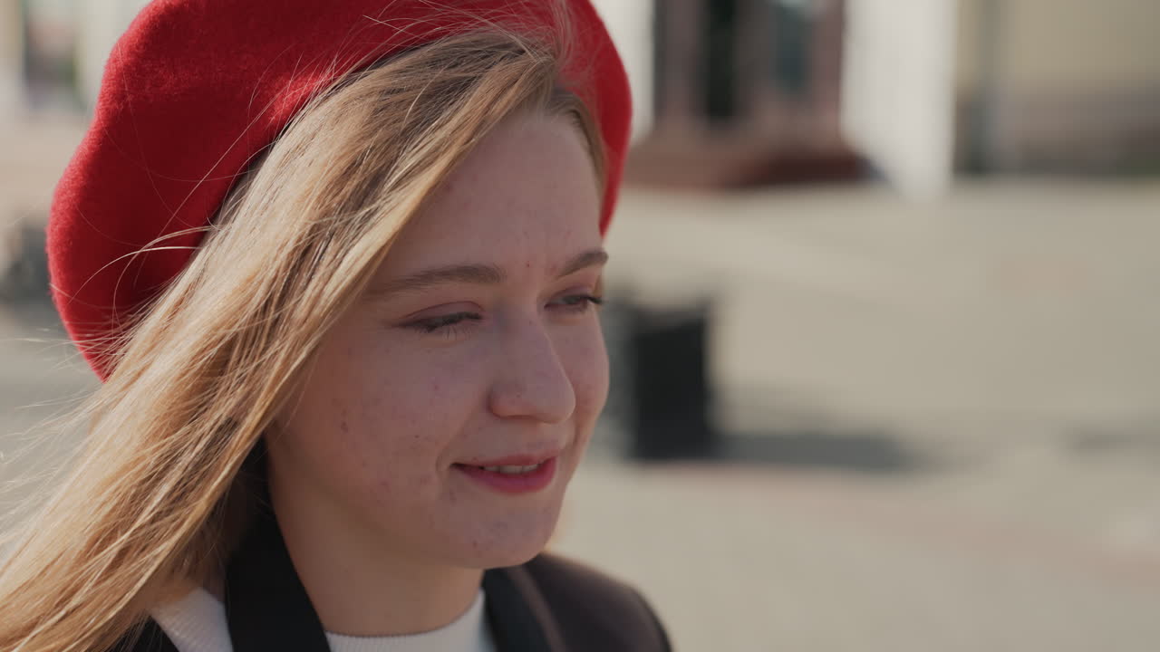 Ung hvid kvinde med rød baret smiler i telefonen på en bænk, afslappet bypause med blødt sollys, hyggelig frakke og nem samtale, blid brise gennem håret og eftertænksomt udtryk.