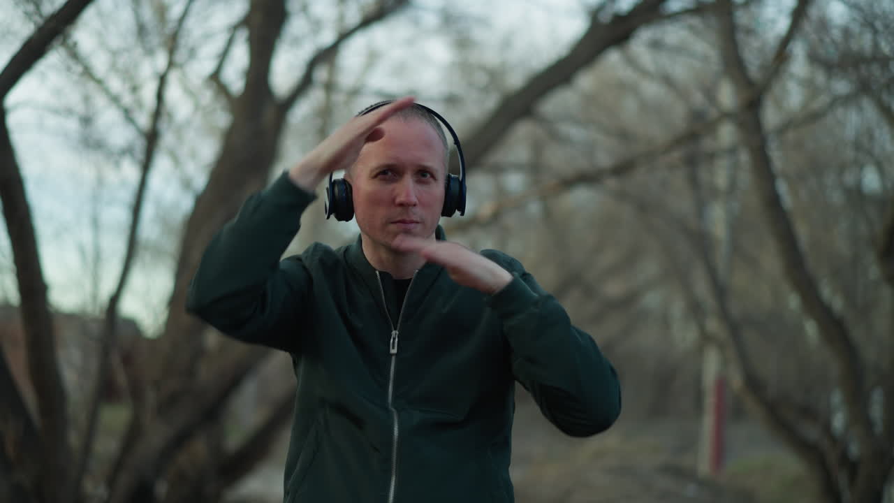 A man in a green jacket and headphones dances with expressive hand movements around his face in a serene forest