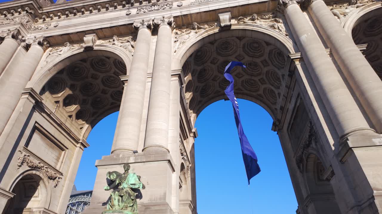 Wide shot of NATO flag waving under Cinquantenaire Arch in Brussels, Belgium, with clear blue sky. Editorial footage capturing political symbolism and European landmark.