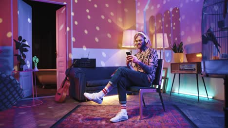 young stylish caucasian man with long hair in headphones sitting on a chair and using smartphone in a retro party at home