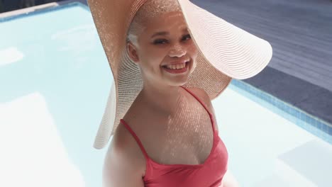 portrait of happy biracial woman with sun hat at swimming pool in slow motion