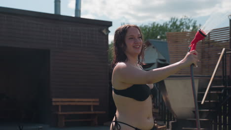 sexy woman in black bikini sprays refreshing water from hose onto herself in sunny backyard grass, droplets catching bright light as she laughs and enjoys playful summer moment of carefree fun