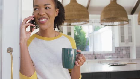 Mujer-Afroamericana-Sosteniendo-Una-Taza-De-Café-Hablando-Por-Teléfono-Inteligente-En-Casa