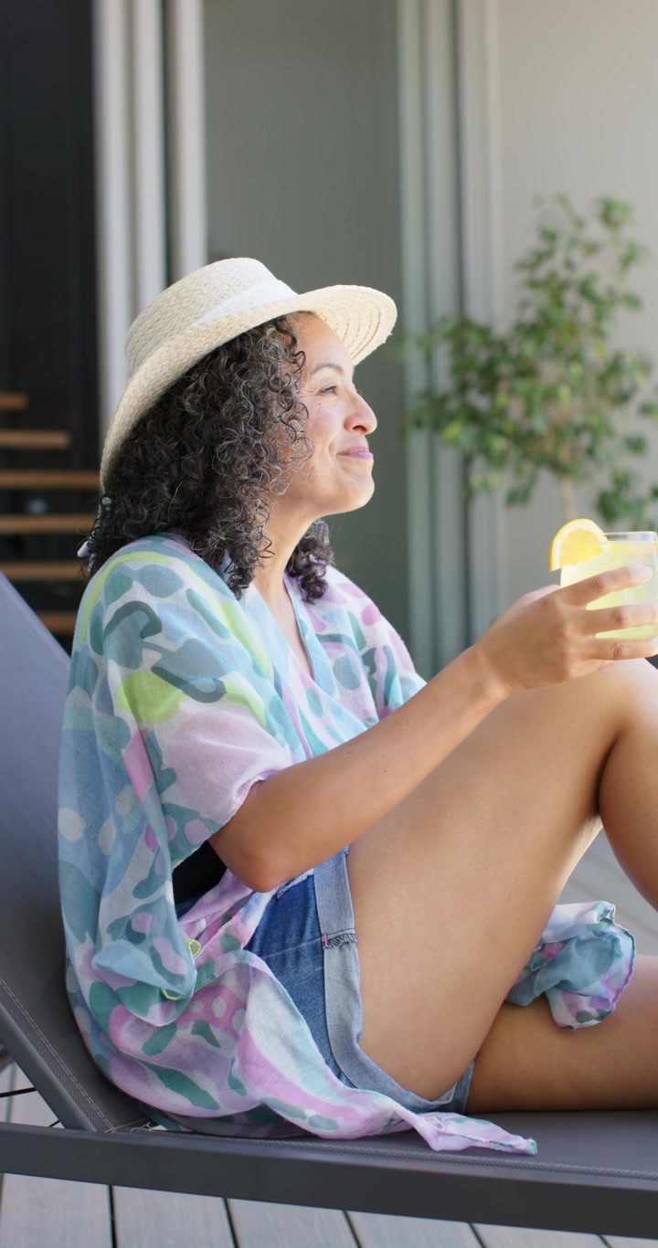 Donna con cappello di paglia che sorseggia un drink, alzando il bicchiere dal ginocchio, mentre si rilassa sul balcone di casa, con una pianta.