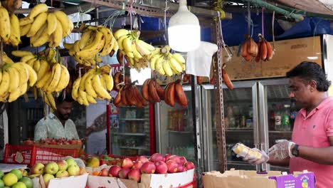 a fruit vendor in a pink shirt prepares and weighs red bananas at a vibrant outdoor market stall in singapore, under bright daylight with steady camera movement