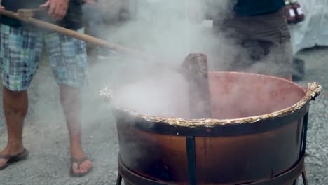 very tight shot of stirring steaming apple butter in a large black cauldron outside