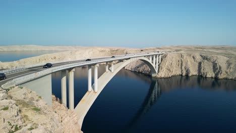 drone liftoff over blue adriatic, arched bridge with traffic and white, stoney island pag, croatia