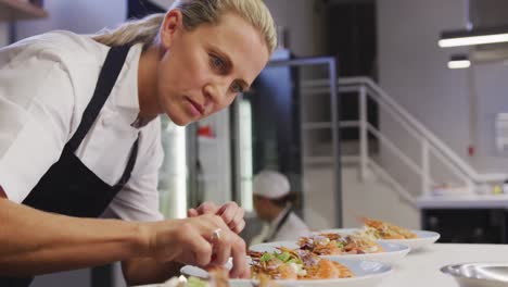 Caucasian-female-chef-wearing-chefs-whites-in-a-restaurant-kitchen,putting-food-on-a-plate