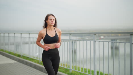 woman running beside iron fence with scenic view of river and bridge in background, showcasing determination and healthy lifestyle in an outdoor urban environment