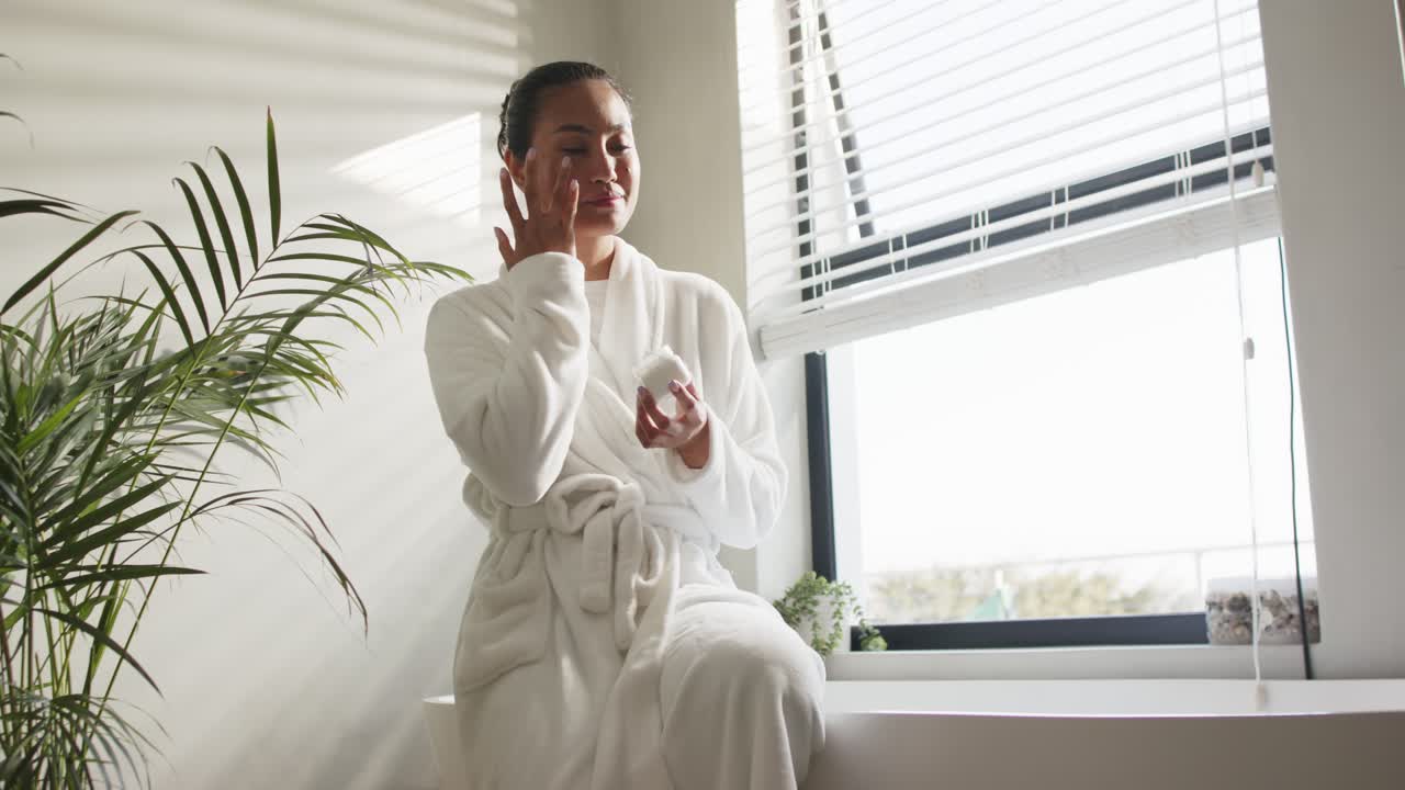 Entering windowsill seat Asian mid adult woman scooping cream and rubbing moisturizer on cheeks