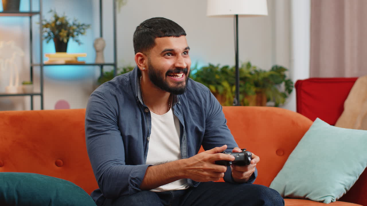 Indian man using joystick controller playing video console winning online fun game sits on home sofa
