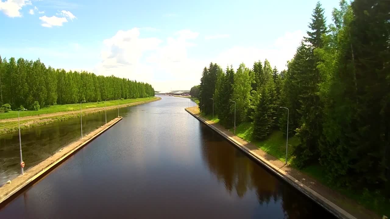 Dronekuva kanavasta Aulangon luonnonsuojelualueella Hämeenlinnassa Suomessa, suoralla vesiväylällä, puiden reunustamilla kävelyteillä ja tyynellä heijastavalla pinnalla sinisen taivaan alla.