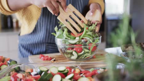 Midsection-of-african-american-woman-in-apron-tossing-salad-in-kitchen,-in-slow-motion