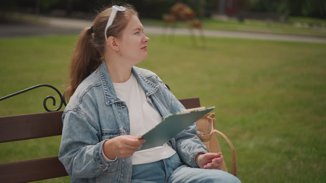 Caucasian lady reviewing notes outside. Female with glasses designing on park bench. Young woman of Caucasian descent pauses thoughtfully with clipboard and glasses outdoors in casual setting