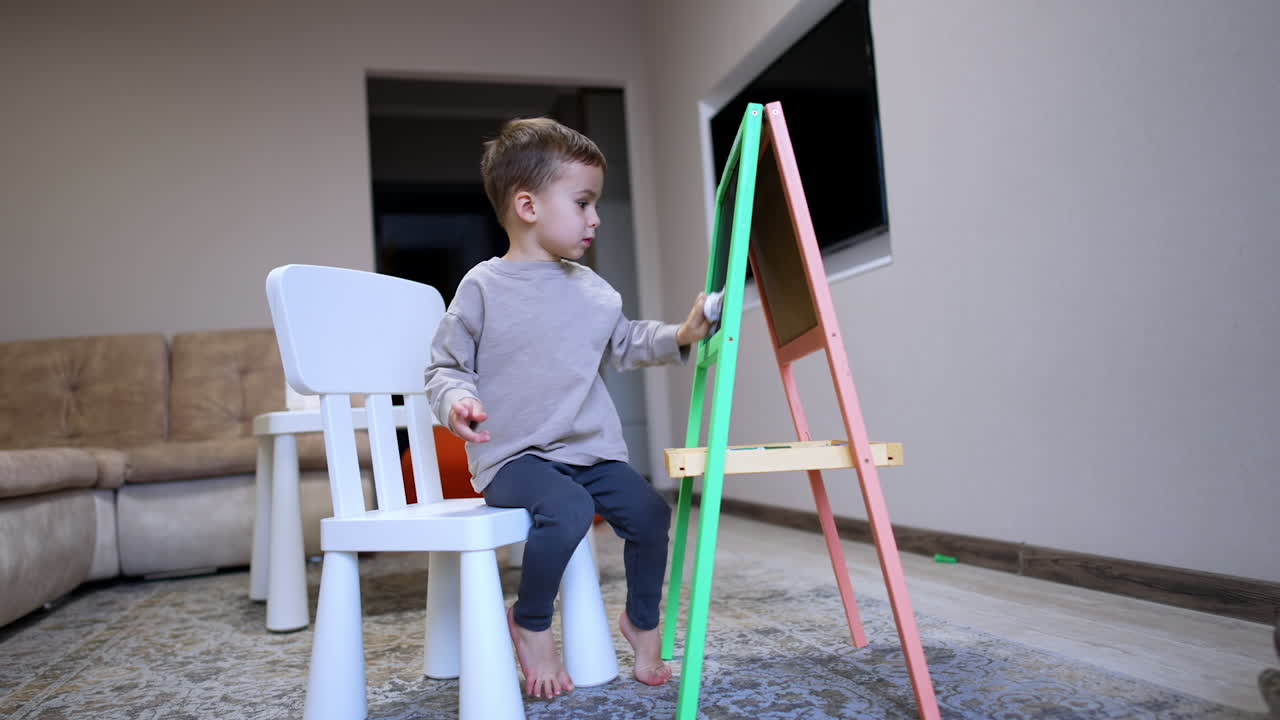 Child painting at easel in living room. Young boy seated on a chair, focused on painting at an easel in a cozy living room while barefoot