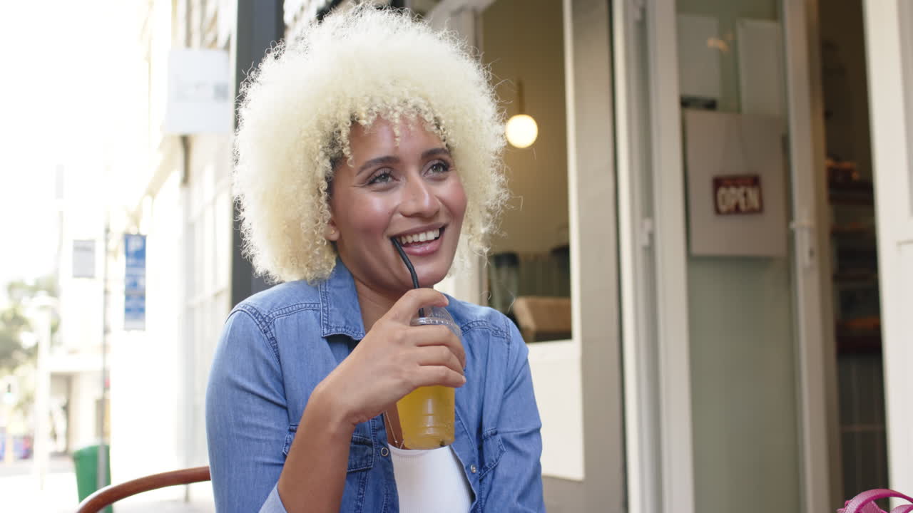 Young biracial woman with curly blonde hair enjoys a drink at an outdoor cafe