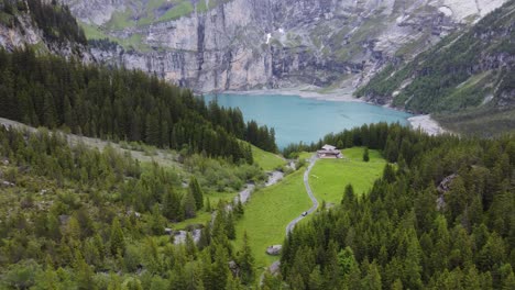 vista aérea coche acercándose desde una granja de cabaña de madera en un verde prado alpino rodeado de montañas alpinas, pinos con vistas al glaciar azul turquesa del lago oeschinensee en kandersteg, suiza