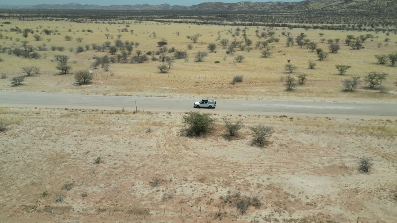 Vista aerea da drone nel deserto della Namibia, con volo sopra una strada stretta che segue un SUV con sabbia e alberi verdi ai lati e montagne all'orizzonte in una giornata di sole con un'auto che sorpassa
