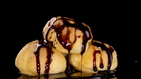 golden cream puffs topped with glossy chocolate sauce are arranged against a black backdrop, with dramatic studio lighting and a static camera angle