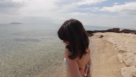 a brunette girl wearing swimwear and smiling at the camera on a sandy beach in the mediterranean on a sunny day