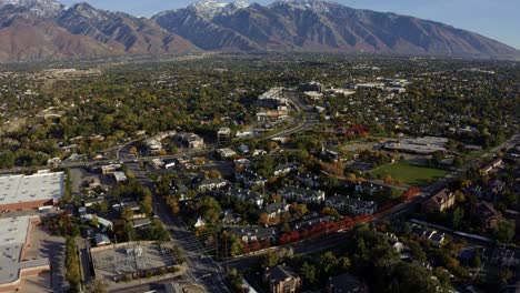 Inclinar-Hacia-Arriba-El-Paisaje-Aéreo-Descendente-De-Drones-Que-Revela-Las-Impresionantes-Montañas-Rocosas-Cubiertas-De-Nieve-De-Utah-Con-El-Condado-De-Salt-Lake-Debajo-Lleno-De-Edificios-Y-árboles-Coloridos-En-Un-Cálido-Y-Soleado-Día-De-Otoño