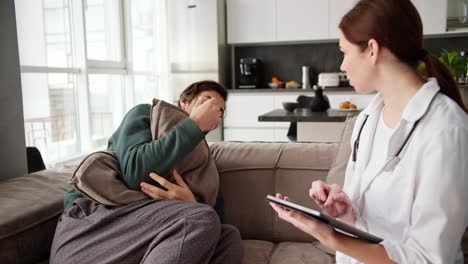 A-tired-and-sick-brunette-man-in-a-green-sweater-hugs-a-pillow-and-sitting-on-the-sofa-tells-a-brunette-girl-to-a-doctor-in-a-white-coat-about-his-symptoms-and-the-girl-writes-them-down-on-her-tablet-during-a-home-examination-in-a-modern-apartment