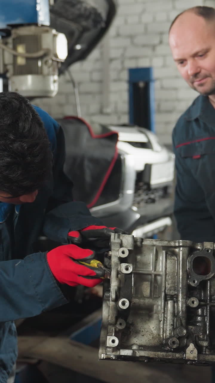dos mecánicos en uniformes azules trabajando en un motor en un taller de automóviles, uno raspando el motor mientras el otro suelta los pernos, rodeados de herramientas y piezas de automóviles