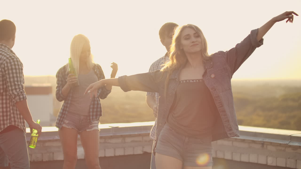la joven hermosa está bailando en el techo con sus cuatro amigas que beben cerveza en la fiesta. ella sonríe y disfruta del tiempo en pantalones cortos y una chaqueta de denim ligera en la noche de verano.