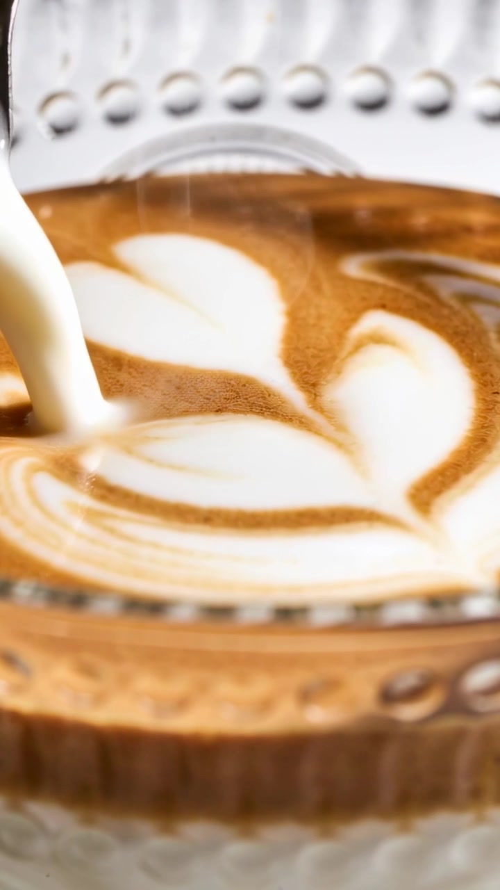 Video verticale: Lattiera che si inclina creando un cuore di latte art sopra una tazza in un caffè, con piattino e vassoio.