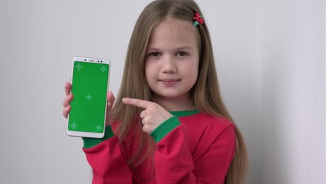 little girl in christmas pajamas shows phone with green screen pointing index finger at mock up cellphone, mobile telephone