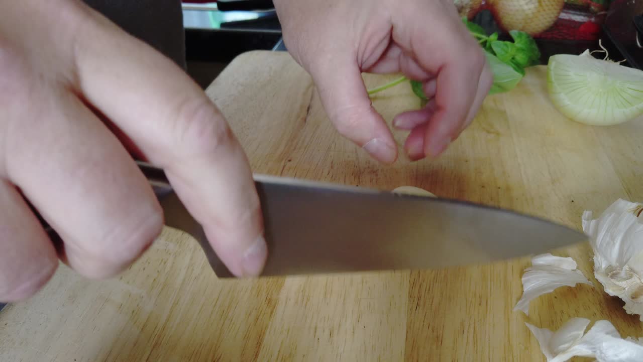 A man crushes and finely chops garlic in a home kitchen on a wooden chopping board