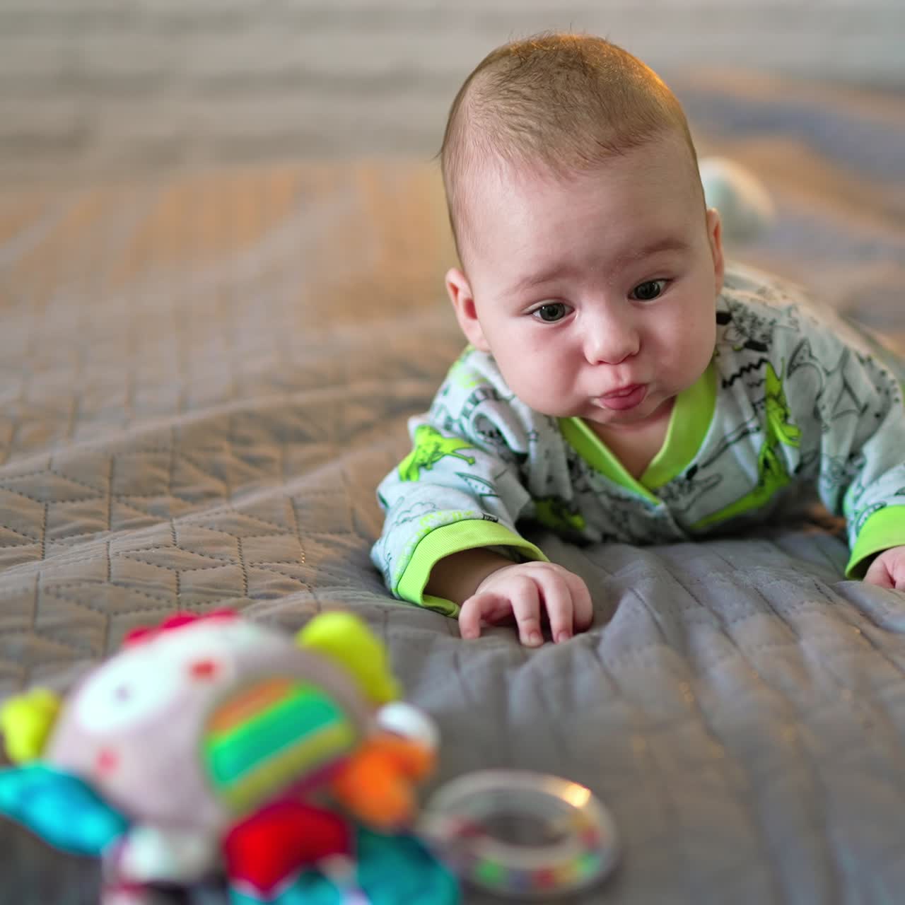 Tiny boy lies on the bed and toy is in front of him. Cute kid moving arms and legs actively, making funny faces. Grey backdrop in blur