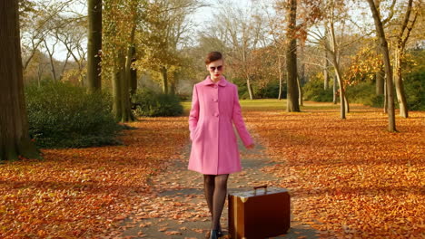 woman in a pink coat walks through an autumn park with a suitcase