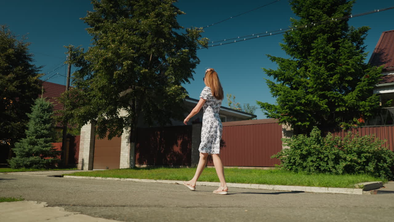 Fair skin woman in white floral dress walks alone on suburban street in daylight, gently scratching head while moving hair behind shoulder, surrounded by trees, houses, and vivid summer greenery