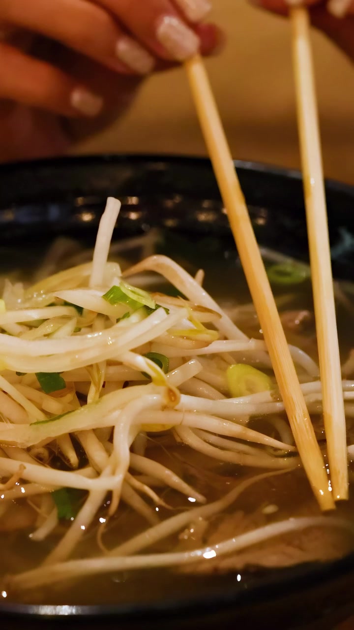 Una persona gusta una ciotola di pho vietnamita, usando le bacchette per sollevare noodles e germogli di soia in un'illuminazione calda.