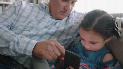 Abuelo-Feliz-Mostrando-A-La-Niña-Cómo-Usar-Un-Teléfono-Inteligente-Enseñando-A-Su-Nieta-Curiosa-Tecnología-Moderna-Niño-Inteligente-Aprendiendo-Teléfono-Móvil-Sentado-Con-El-Abuelo-En-El-Sofá