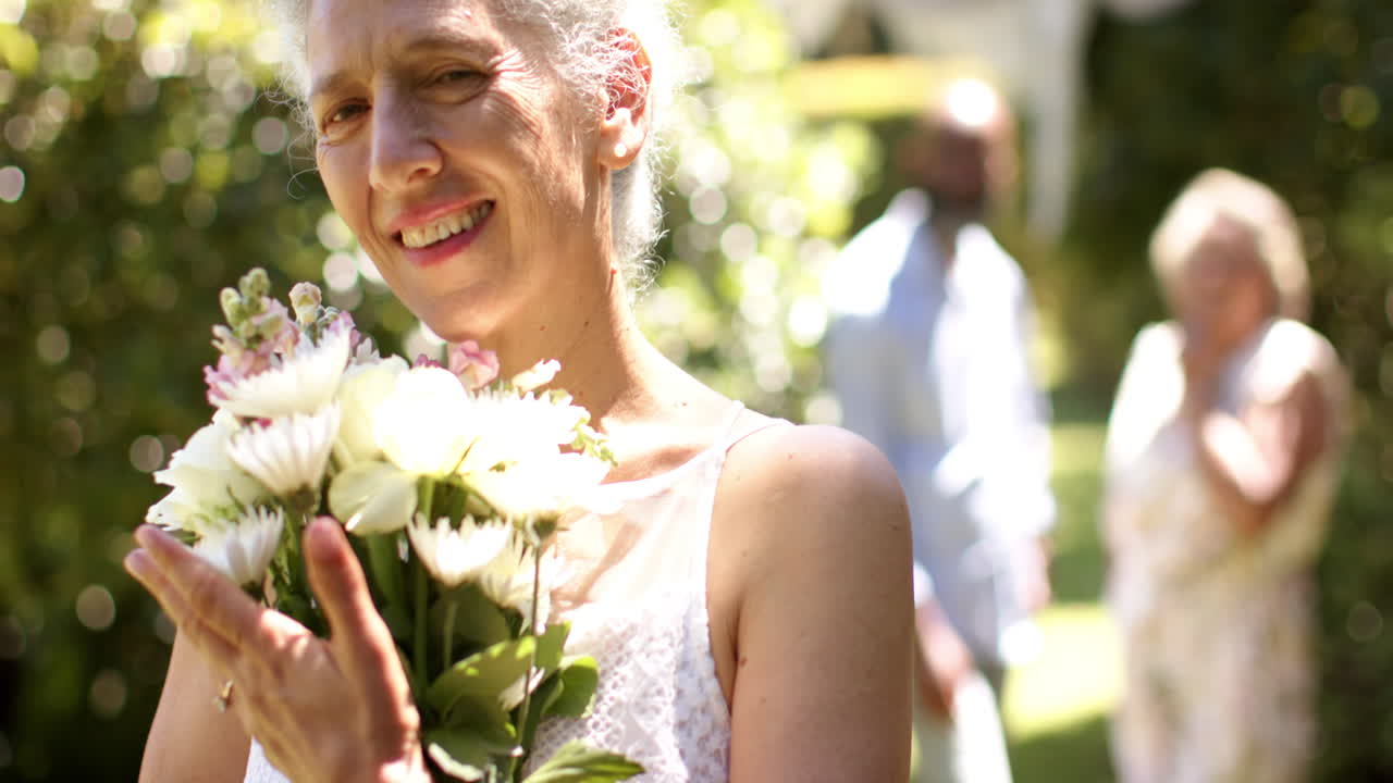 Smiling senior bride holding bouquet of flowers at outdoor wedding with friends in background