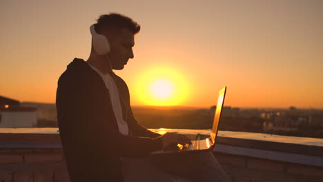 a man in headphones on the roof relaxes working remotely enjoying life despite a handsome kind of sipping beer and types on the keyboard. trade on the stock exchange using a laptop and enjoying the beautiful view