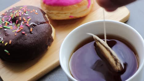 close up hand of woman dripping tea bag in a tea cup with delicious donuts on wooden cutting board.